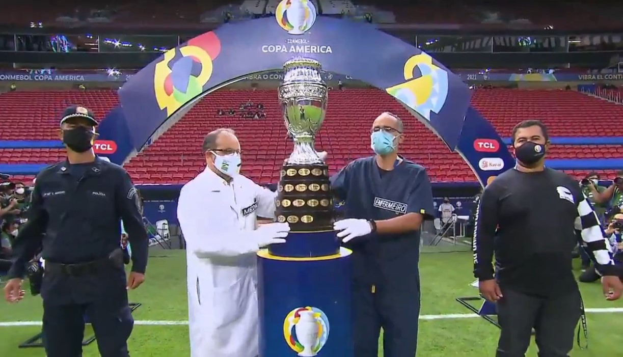 Un médico y un enfermero entran al estadio en Brasilia, en la inauguración de la Copa América, sosteniendo el trofeo. Foto: Twitter / Conmebol